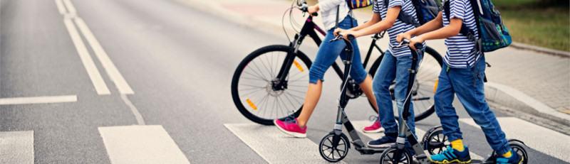 Kinder mit Tretrollern und Fahrrad überqueren einen Zebrastreifen.